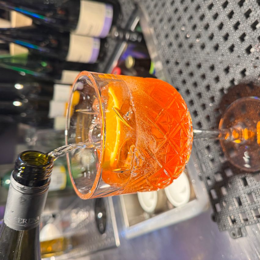 Eine Flasche Sekt wird in ein dekoratives Glas mit Orangensaft und Eis gegossen. Das Glas steht auf einer Metalloberfläche mit einem Netzdesign.