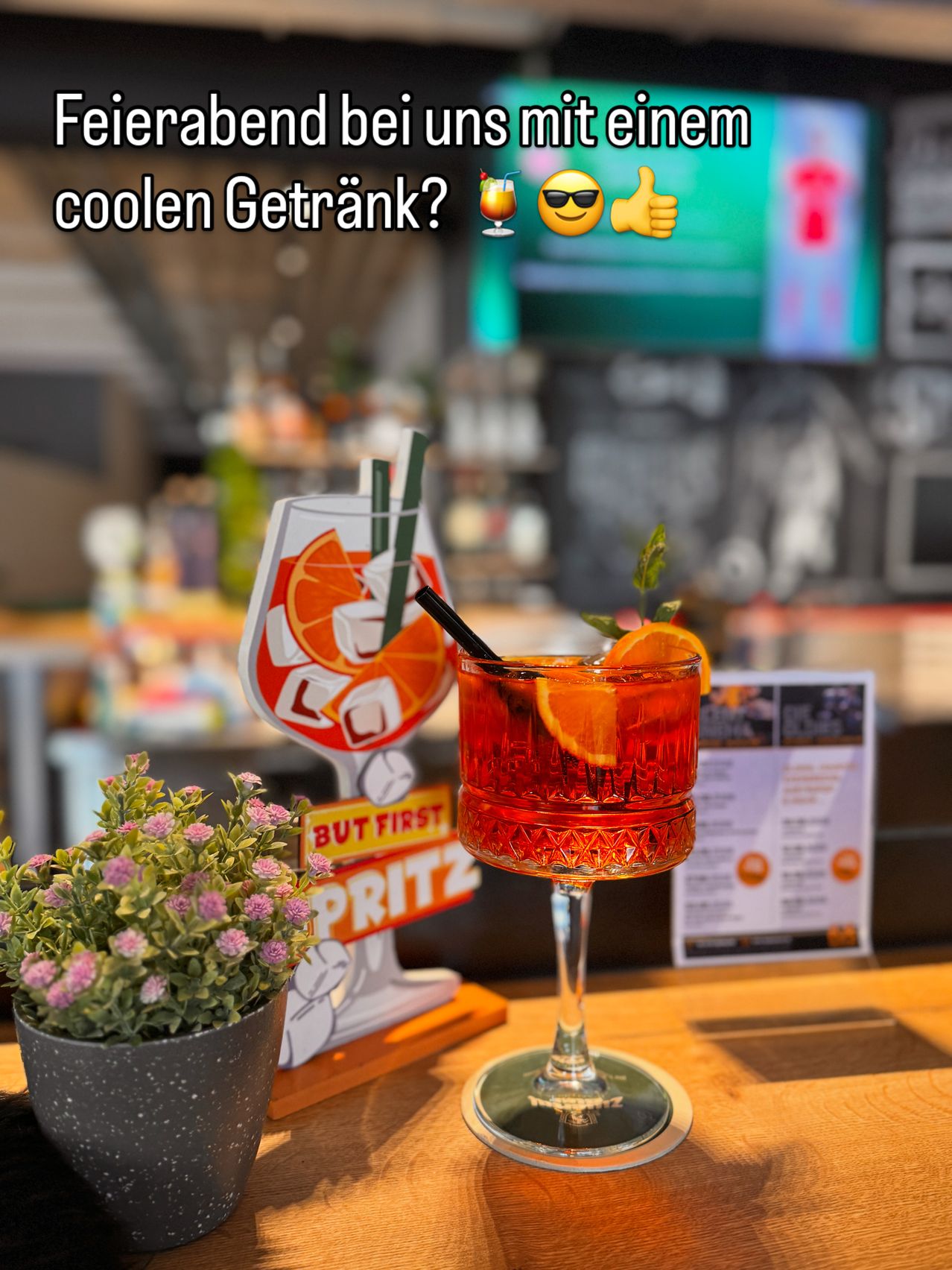 Ein Cocktailglas mit einem roten Getränk, garniert mit einer Orangenscheibe und einem Strohhalm, steht auf einem Holztisch in einer Bar. Ein Topf mit Pflanzen und ein Schild mit der Aufschrift 'Aber zuerst, Spritz' sind in der Nähe.