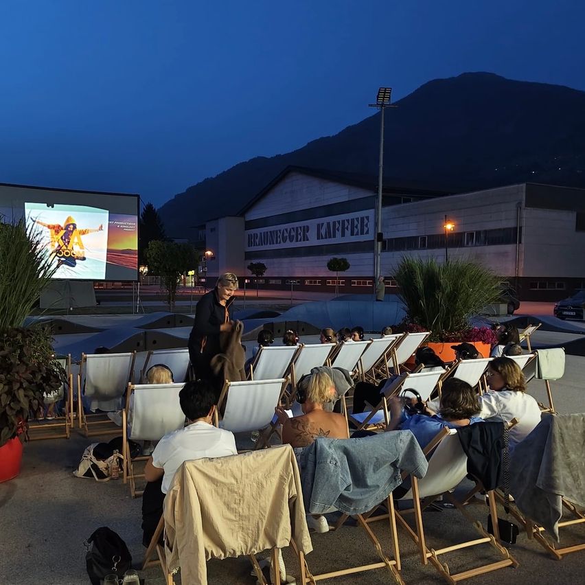 Eine Gruppe von Menschen sitzt in Stühlen und schaut sich einen Film auf einer Leinwand im Freien bei Nacht an. Eine Frau steht vor ihnen, und im Hintergrund befindet sich ein Gebäude mit einem Schild, das 'BRAUNEGGER KAFFEE' besagt.