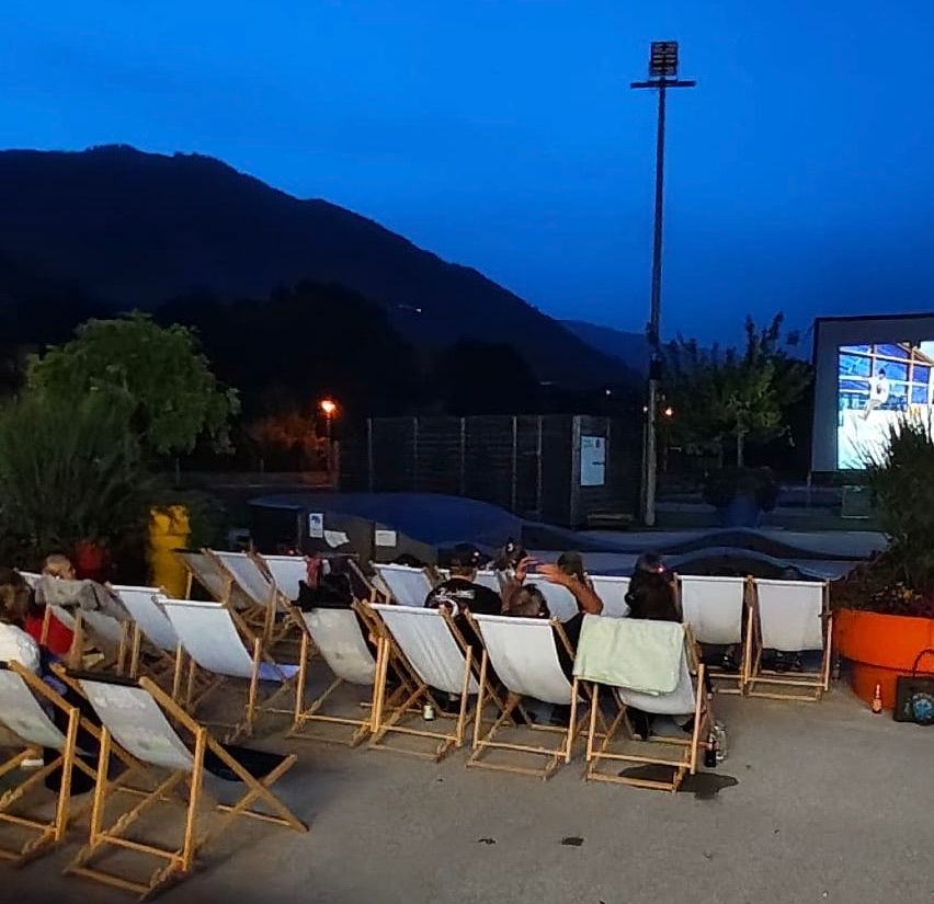Eine Gruppe von Menschen sitzt auf weißen Klappstühlen in einem Außenbereich und schaut sich einen Film auf einer großen Leinwand an. Die Umgebung ist in der Dämmerung, mit Bergen im Hintergrund und einem klaren blauen Himmel.
