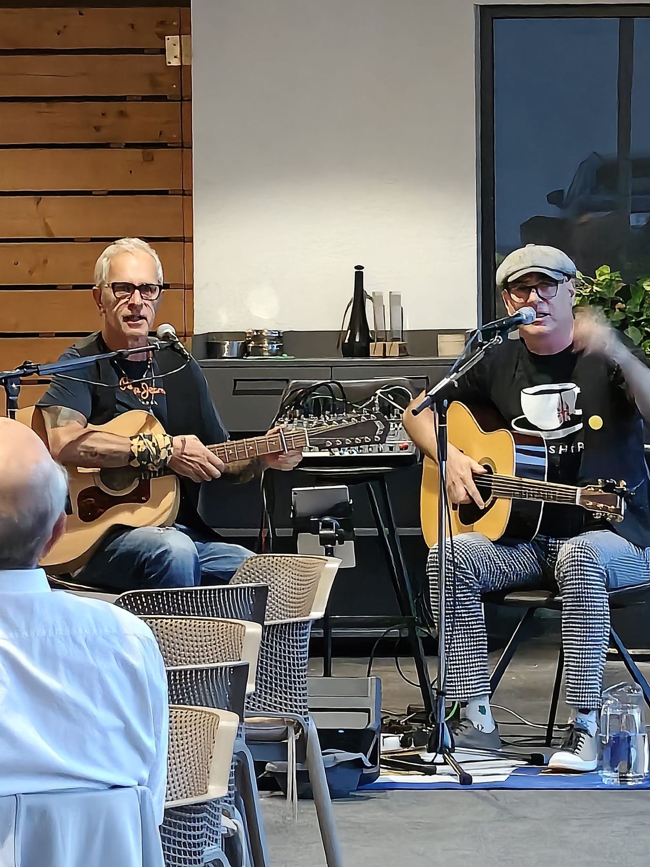 Zwei Männer spielen Gitarren auf der Bühne mit Mikrofonen vor ihnen. Einer trägt einen Hut und ein Kaffeetassen-Shirt, der andere hat ein Tattoo auf seinem Arm. Ein Mann beobachtet vom Publikum aus.