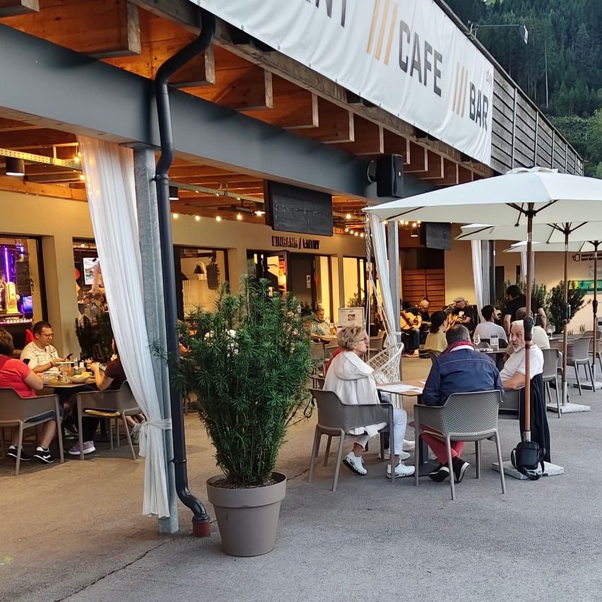 Sitzbereich im Freien in einem Café mit Gästen unter Schirmen, einem Topfpflanzen und einem Banner mit der Aufschrift Cafe Bar.