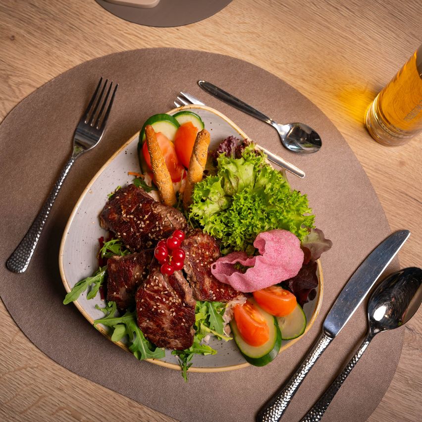Ein Teller mit Steak, gemischtem Gemüse und Salat, begleitet von Besteck und einem Glas Bier auf einem Holztisch.
