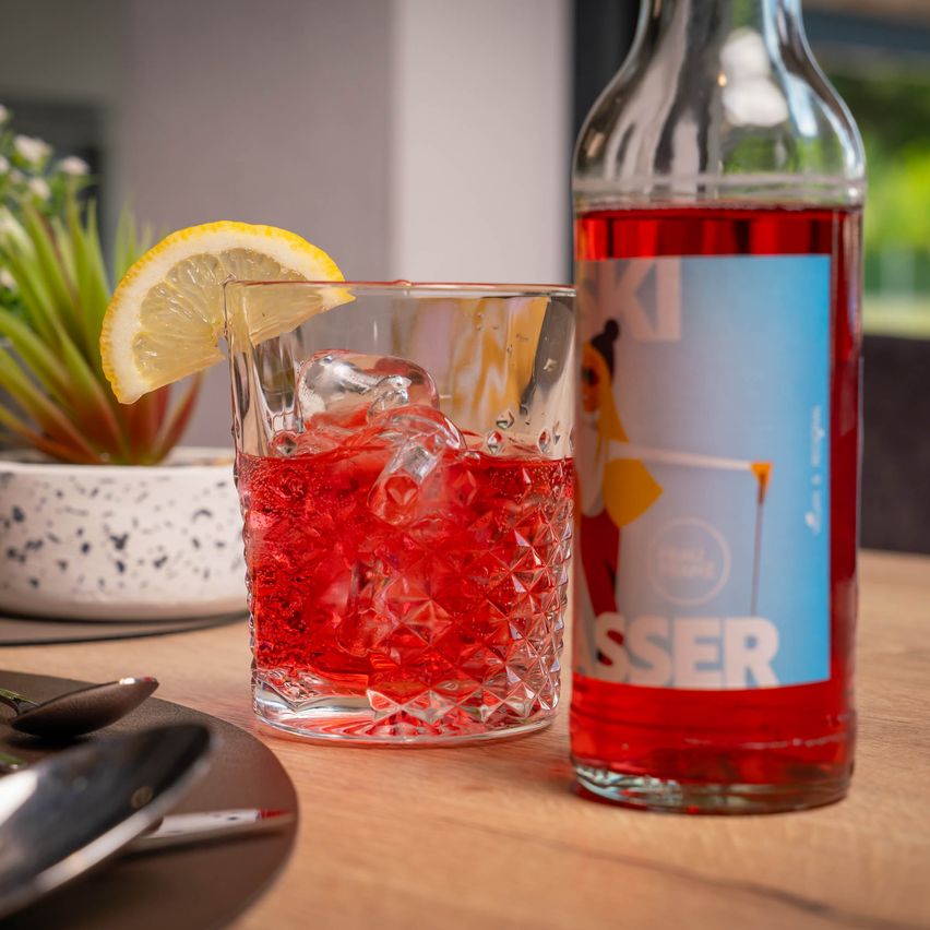 Ein Glas roter Cocktail mit Eiswürfeln und einer Zitronenscheibe auf einem Holztisch neben einer Flasche roter Flüssigkeit.