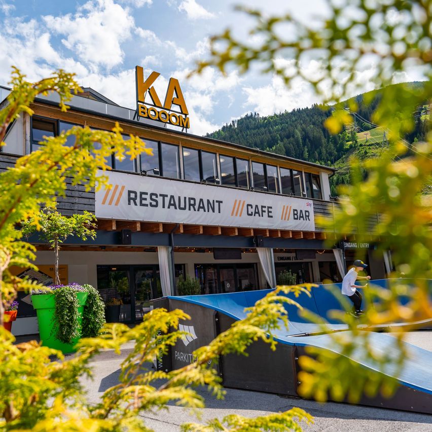Ein Gebäude mit einem Schild, das KA BOOM oben drauf hat. Es hat ein Banner darunter, das Restaurant, Café und Bar sagt. Ein Junge skateboardet auf einer Rampe vor dem Gebäude.
