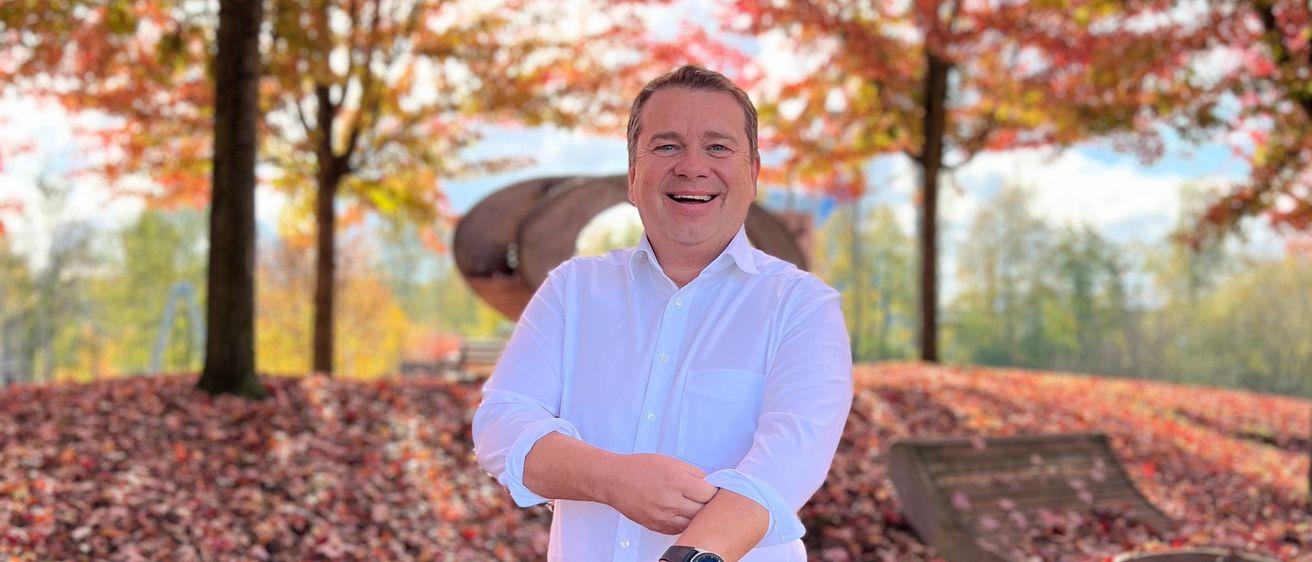 Ein Mann in einem weißen Hemd und blauen Jeans steht in einem Park mit bunten Blättern, lächelt und hält sich mit den Händen an der Taille. Hinter ihm stehen Bäume mit Herbstblättern.