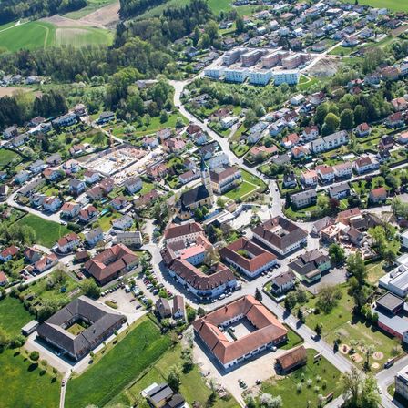 Eine Luftaufnahme einer Stadt mit vielen Gebäuden und Häusern, umgeben von grünen Feldern, Bäumen und einer Straße. Auf der linken Seite befindet sich eine Baustelle.