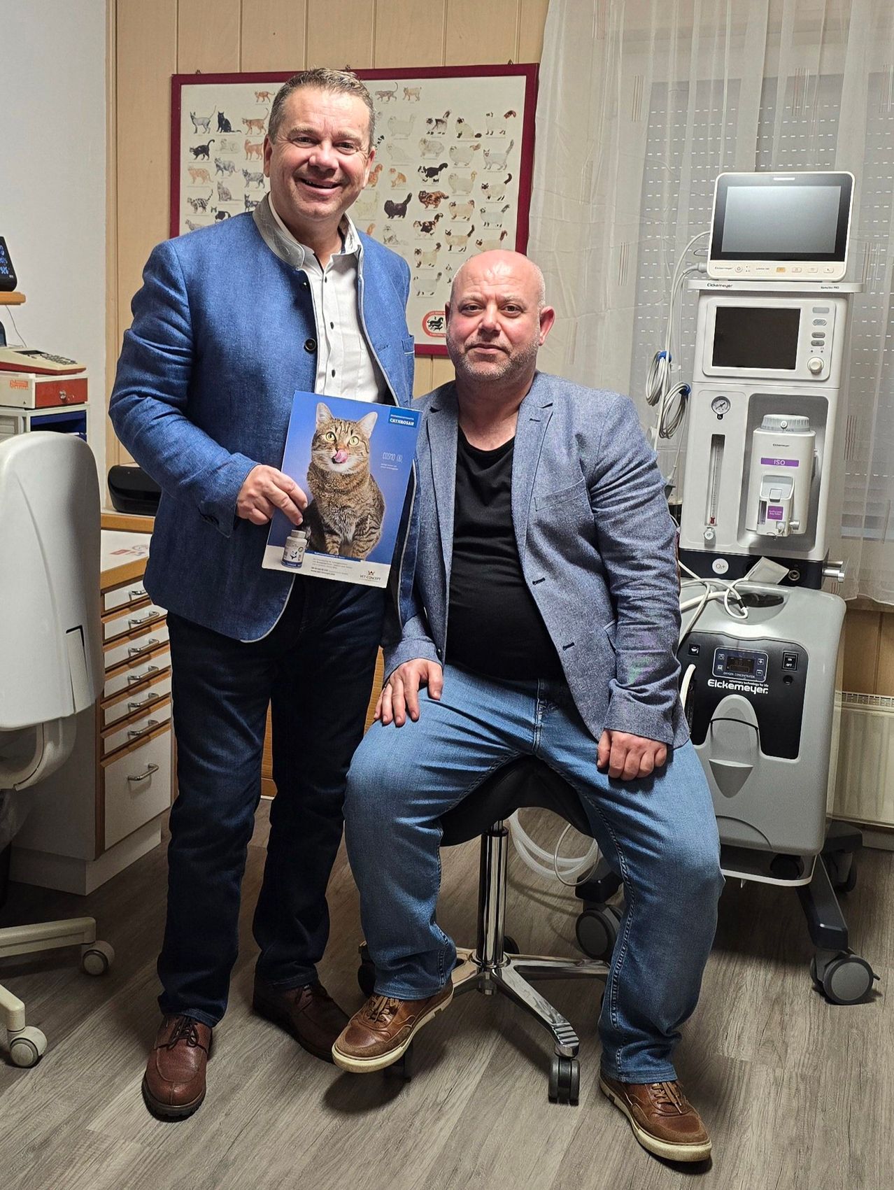 Zwei Männer posieren für ein Foto in einem Raum mit medizinischen Geräten. Ein Mann hält ein Poster einer Katze.