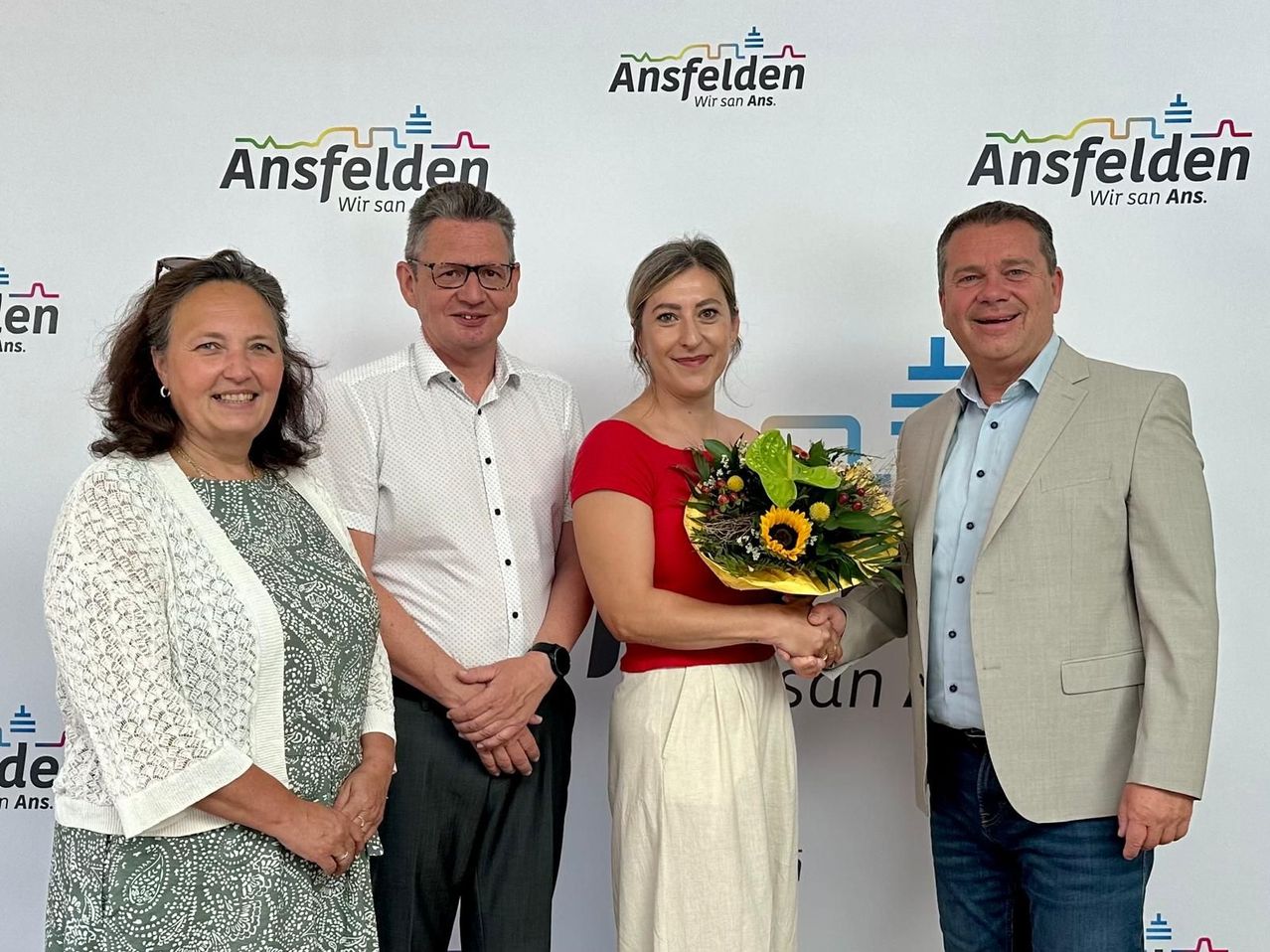 Vier Personen stehen vor einer Wand mit Ansfelden-Markenzeichen. Eine Frau in Rot hält einen Blumenstrauß, lächelt, während die anderen zuschauen.