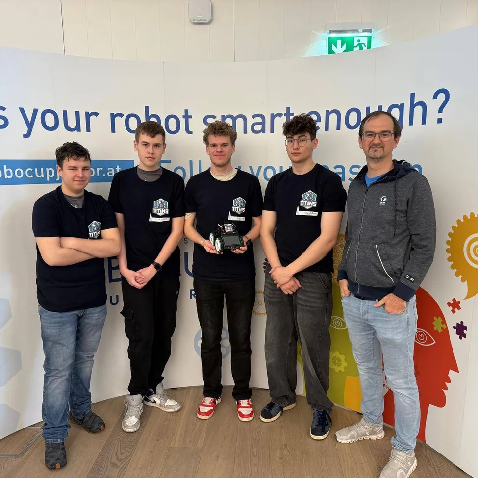 Fünf junge Männer stehen vor einer farbenfrohen Wand, drei halten einen Roboter und tragen schwarze T-Shirts mit Logos. Der Mann rechts trägt eine graue Kapuzenjacke.