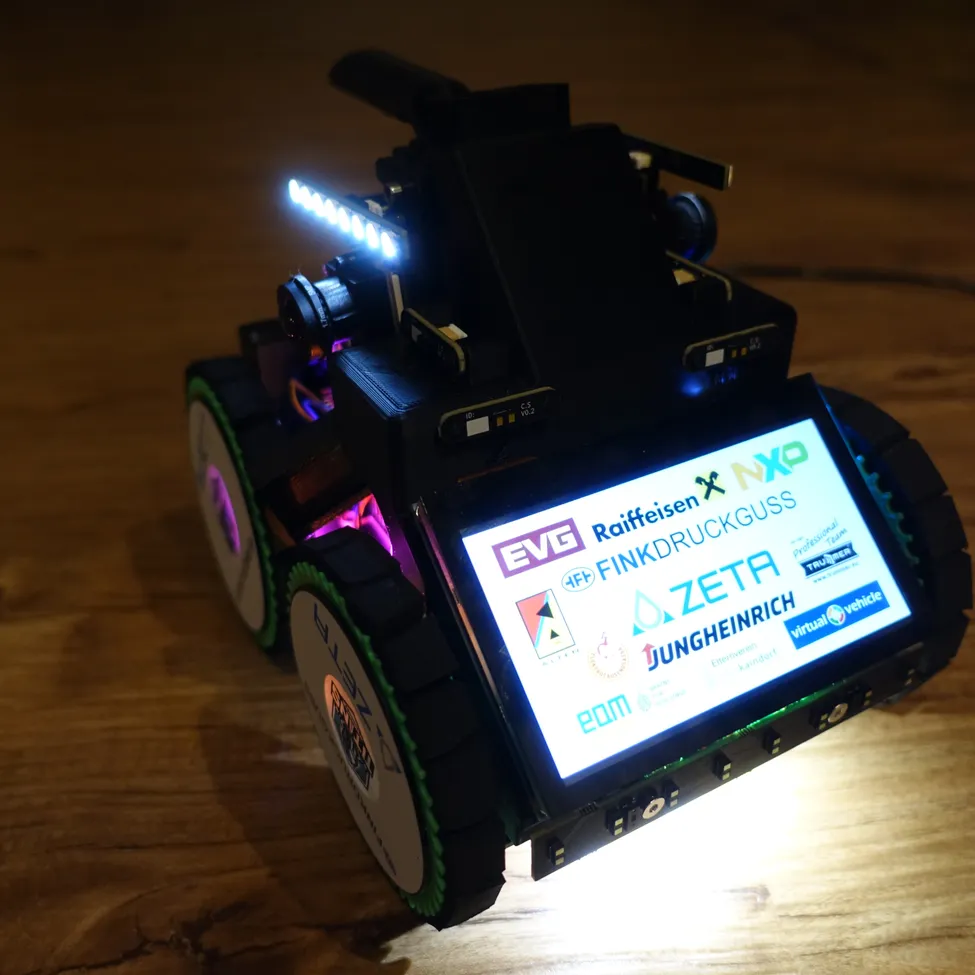 Ein Roboterfahrzeug mit beleuchtetem Bildschirm zeigt Logos von EVG, Raiffeisen und anderen. Es hat eine montierte Kamera und blaue und lila Lichter.