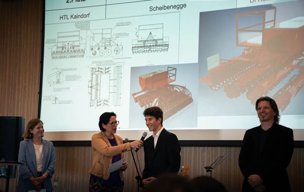 Eine Frau mit Brille spricht in ein Mikrofon und hält ein Papier in der Hand, während sie einen Mann in einem schwarzen Anzug interviewt. Ein Beamer hinter ihnen zeigt technische Diagramme und eine rote Maschine.