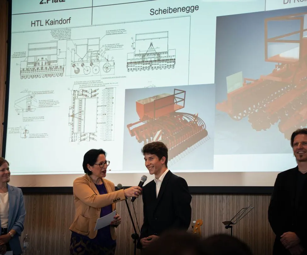 Eine Frau mit Brille spricht in ein Mikrofon und hält ein Papier in der Hand, während sie einen Mann in einem schwarzen Anzug interviewt. Ein Beamer hinter ihnen zeigt technische Diagramme und eine rote Maschine.