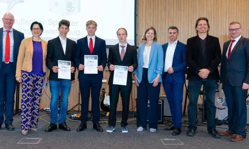 Eine Gruppe von Personen steht gemeinsam für ein Foto, hält Urkunden mit Namen und Nummern in den Händen. Einige tragen Anzüge, und eine Gitarre befindet sich hinter ihnen. Der Hintergrund besteht aus einer Holzwand und einer Projektionsleinwand.