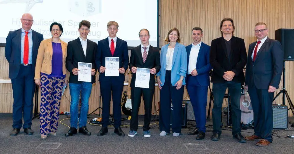 Eine Gruppe von Personen steht gemeinsam für ein Foto, hält Urkunden mit Namen und Nummern in den Händen. Einige tragen Anzüge, und eine Gitarre befindet sich hinter ihnen. Der Hintergrund besteht aus einer Holzwand und einer Projektionsleinwand.