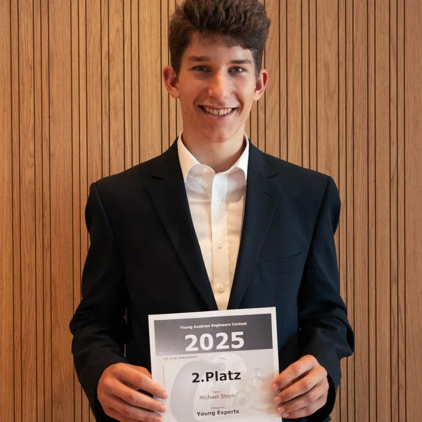 Ein Mann hält ein Zertifikat in den Händen, das 2. Platz und 2025 zeigt. Er lächelt und ist in einem schwarzen Anzug und einem weißen Hemd gekleidet.