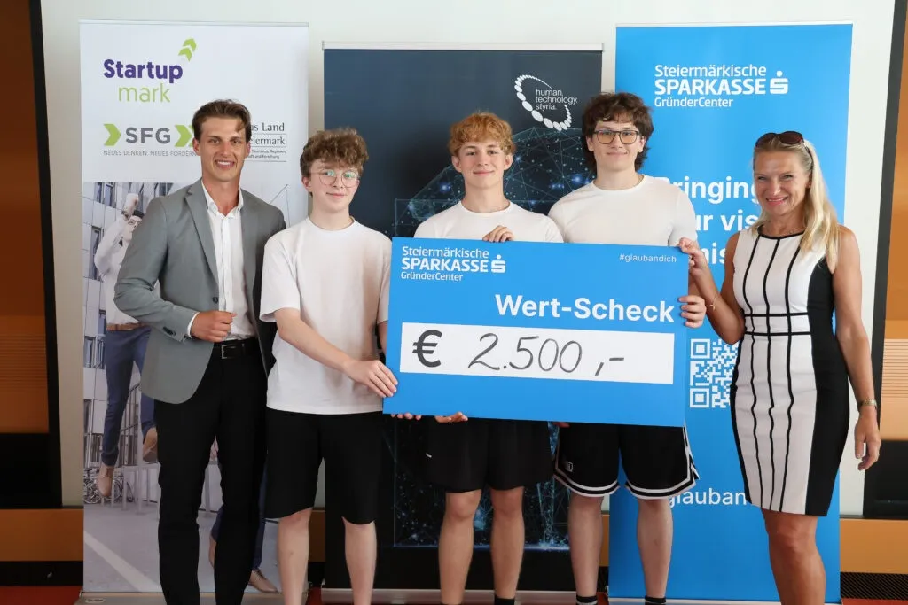 Eine Gruppe junger Menschen, drei Männer und eine Frau, stehen mit einem großen blauen Scheck über 2.500 Euro und halten ihn vor einem großen Plakat.