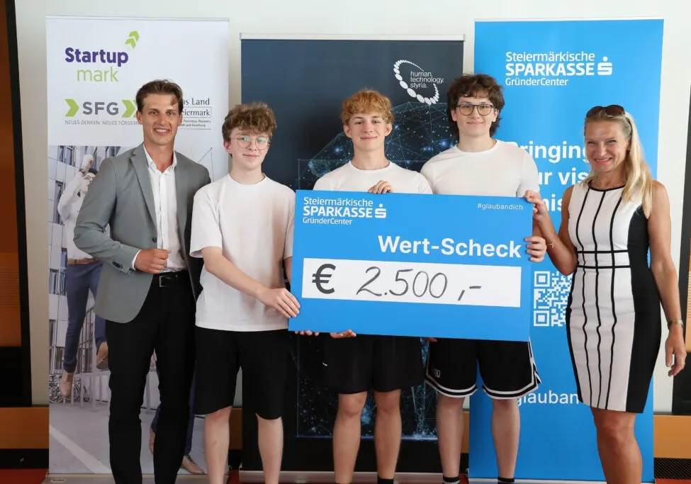 Eine Gruppe junger Menschen, drei Männer und eine Frau, stehen mit einem großen blauen Scheck über 2.500 Euro und halten ihn vor einem großen Plakat.