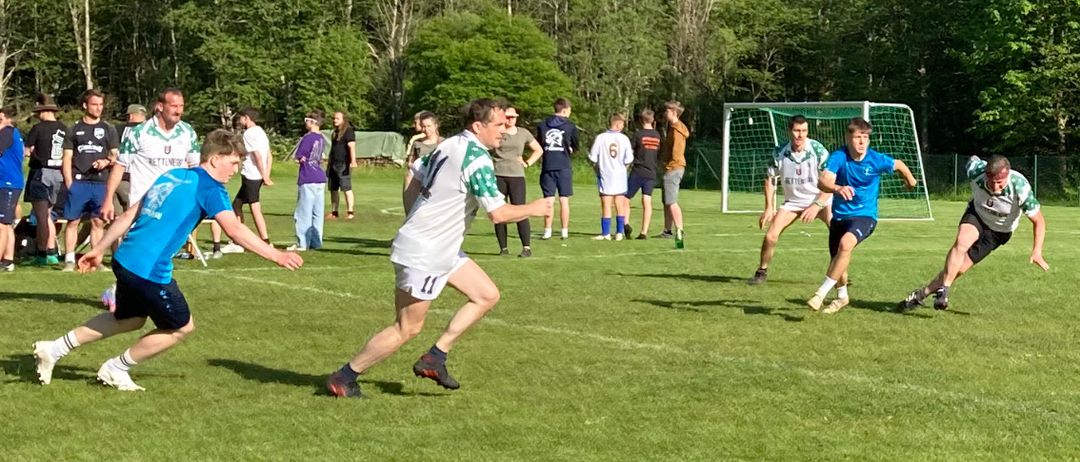 Eine Gruppe von Menschen spielt Fußball auf einem Feld. Ein Mann in einem weißen Trikot läuft auf das Tor zu.