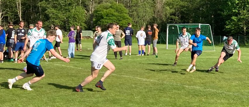 Eine Gruppe von Menschen spielt Fußball auf einem Feld. Ein Mann in einem weißen Trikot läuft auf das Tor zu.
