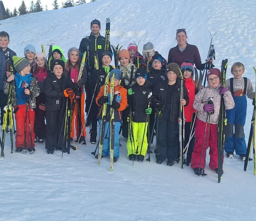 Eine Gruppe von Kindern und Erwachsenen steht im Schnee und hält Skier und Stöcke. Einige tragen Mützen, Handschuhe und Brillen. Bäume und ein verschneiter Hügel sind im Hintergrund.