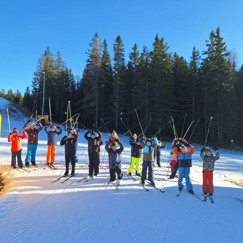 Eine Gruppe von Kindern in Skiausrüstung steht in einer Reihe auf einem verschneiten Hang, hält ihre Skistöcke und schaut nach oben, mit Bäumen im Hintergrund.