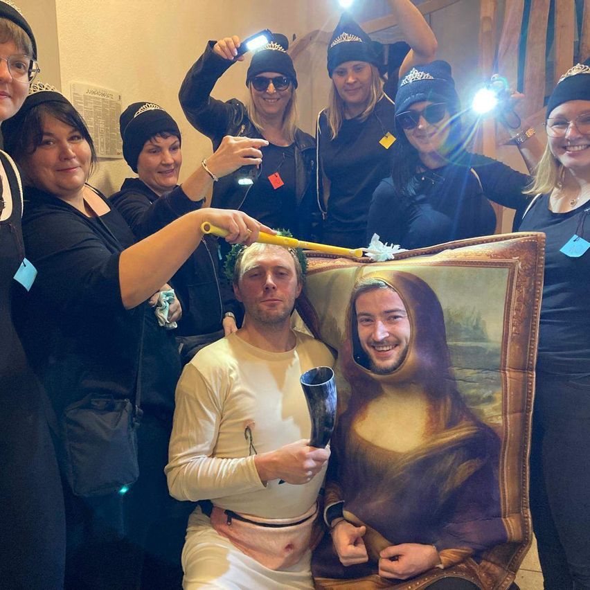 A group of women dressed in black and wearing crowns take a photo with a man dressed as Mona Lisa and another man holding a horn.