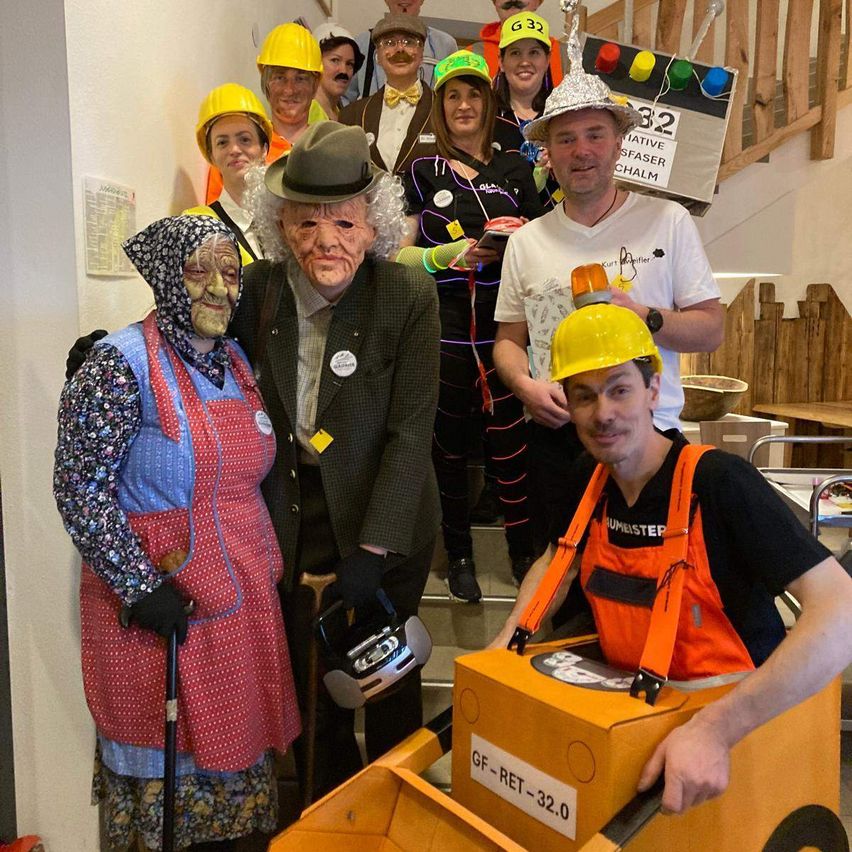A group of people in costumes are standing and smiling for a photo. One person is in an orange excavator costume. Another person is wearing a hat and holding a radio. Some people are wearing yellow hard hats. A person is wearing a mask and holding a cane. A wooden staircase is behind them.