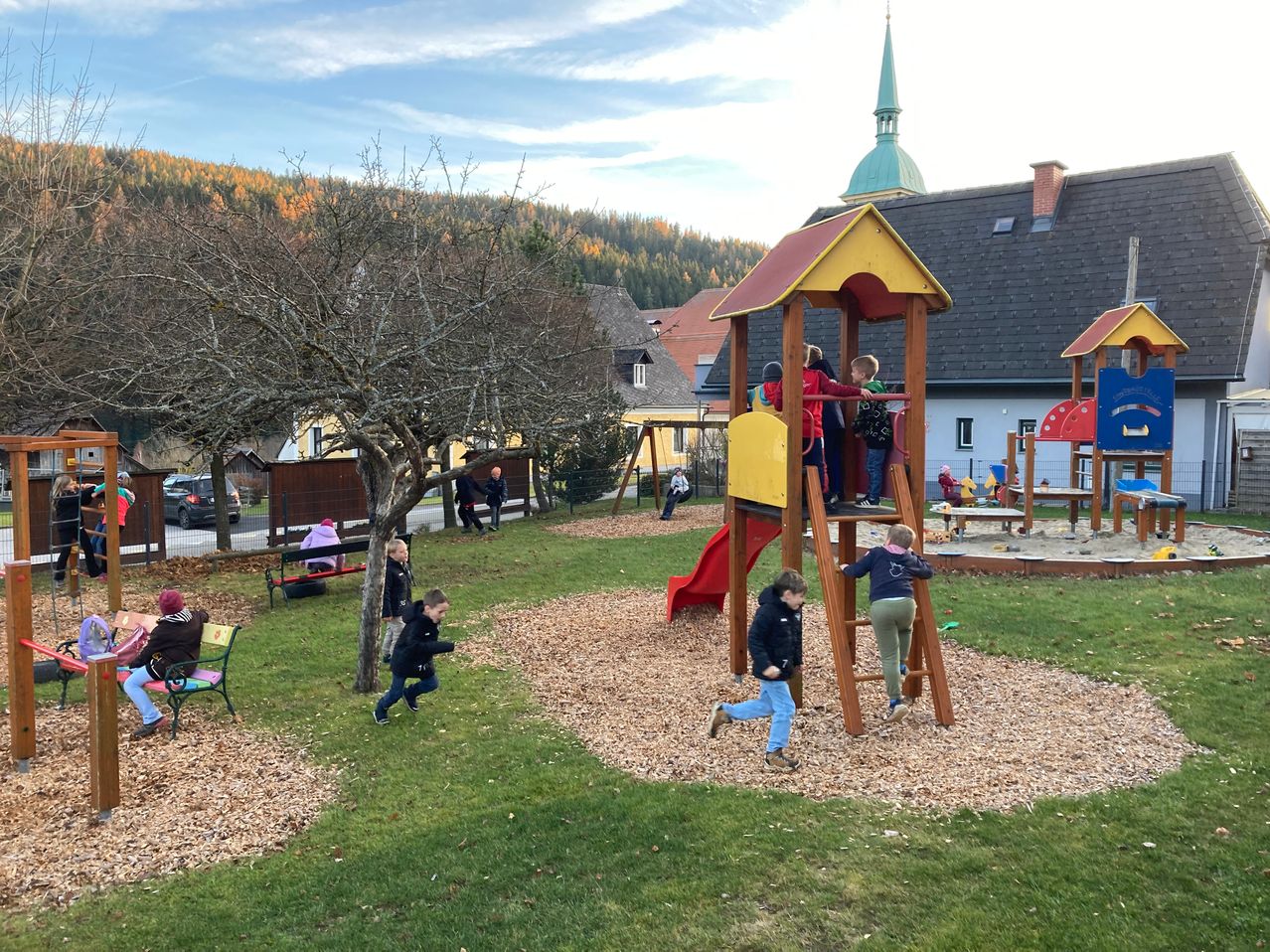 Children play on a playground with a slide and climbing frame, with houses and mountains in the background.