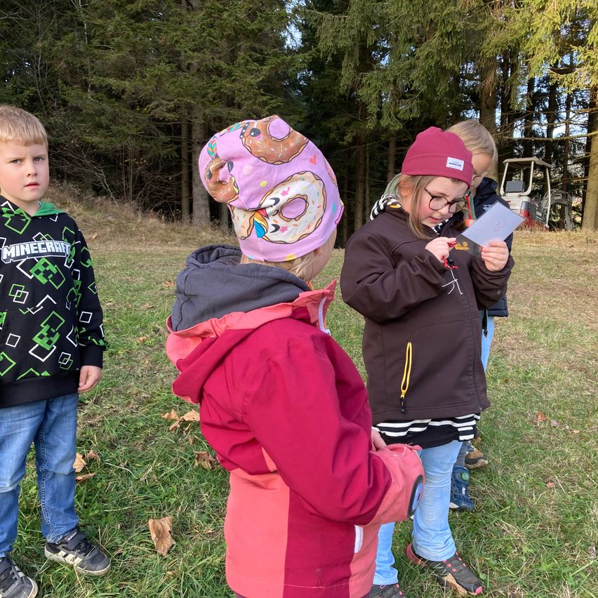A group of kids are in a grassy field, with one girl reading a paper and another looking at something. Behind them are trees and a vehicle.