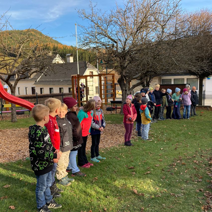 A group of children wearing winter clothing stands in a grassy area near a playground, with trees and a house in the background.