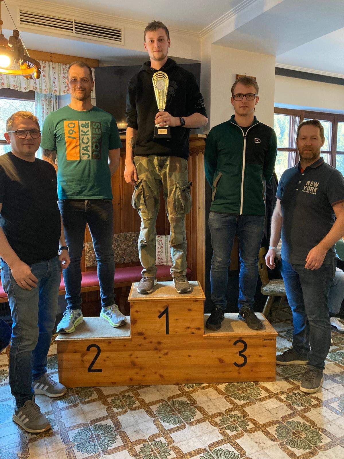 Fünf Männer stehen auf einem Holzpodium mit den Nummern 1, 2 und 3. Ein Mann hält einen Pokal. Sie befinden sich in einem Raum mit Holzboden und einer Wand mit Fenster.