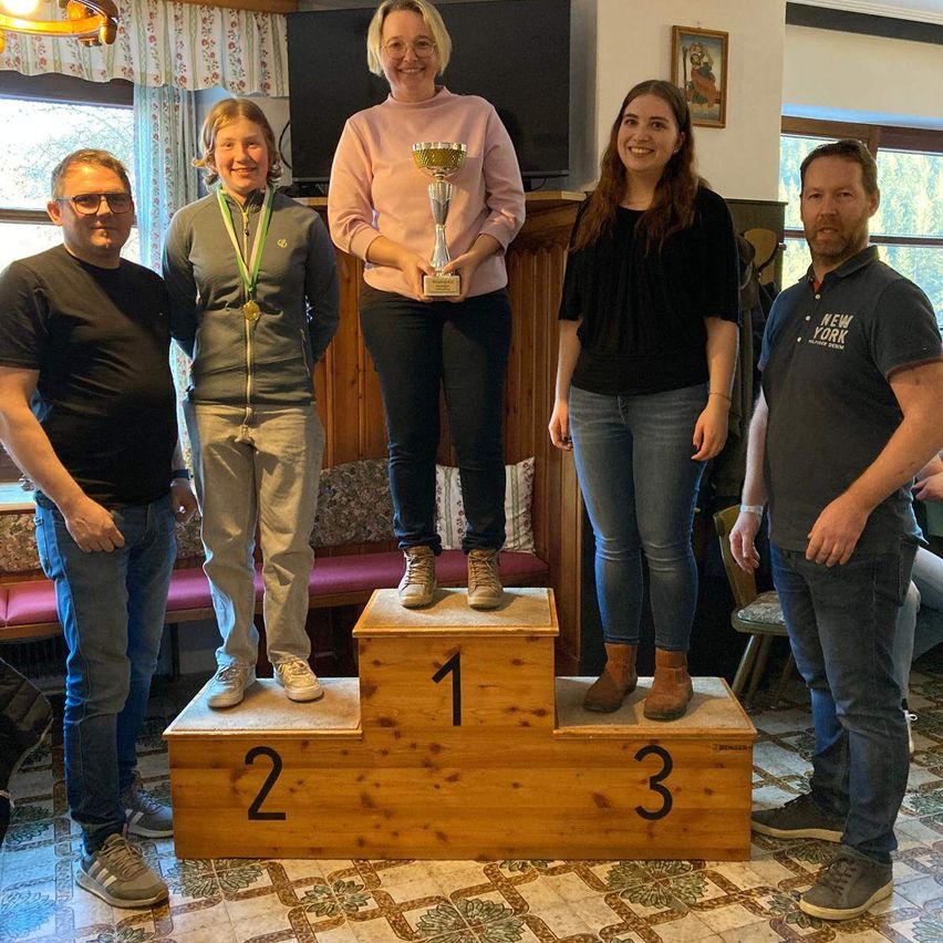 Fünf Personen stehen auf einem Podest mit den Nummern eins bis drei. Eine Frau hält einen Pokal. Die Frau in der Mitte lächelt und schaut in die Kamera. Dahinter steht ein Holzschrank mit einem Fernseher darauf. An der Wand hängt ein gerahmtes Bild.