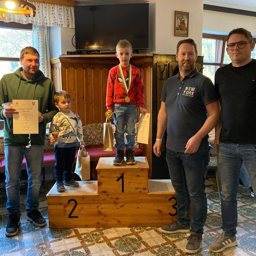 Fünf Männer und Jungen stehen auf einem Podium, zwei von ihnen halten Urkunden. Der Junge in der Mitte trägt eine Medaille. Sie lächeln und scheinen für ein Foto zu posieren. Hinter ihnen steht ein Fernseher, ein Schrank und ein Bilderrahmen.