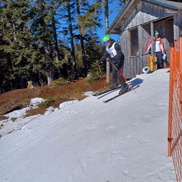 Ein Skifahrer mit grünem Helm fährt einen schneebedeckten Hang hinunter, mit einem Holzgebäude im Hintergrund.