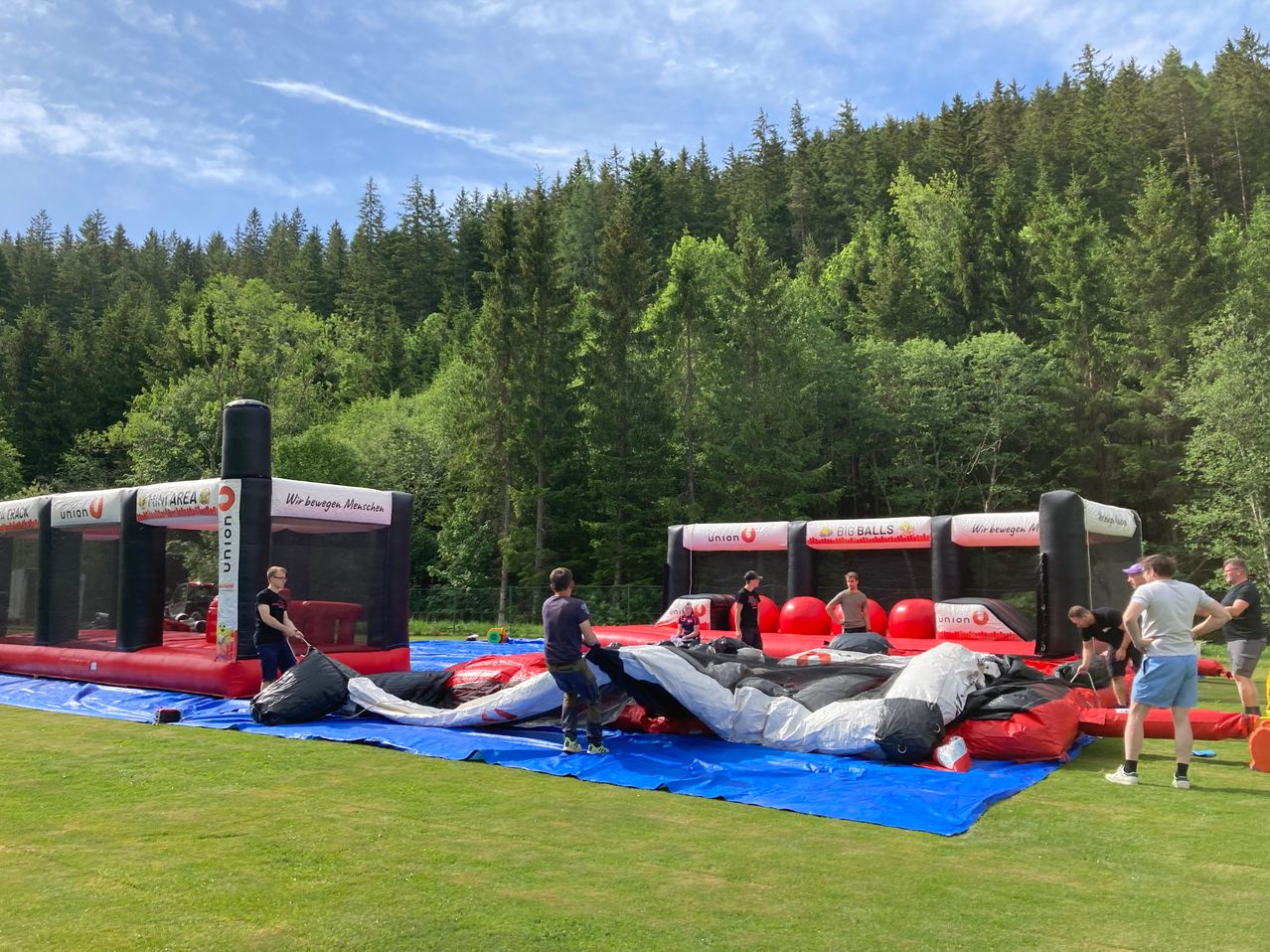 Auf einer Wiese mit Bäumen im Hintergrund werden aufblasbare Hüpfburgen aufgebaut. Einzelpersonen sind aktiv an der Vorbereitung der Ausrüstung für den Gebrauch beteiligt.