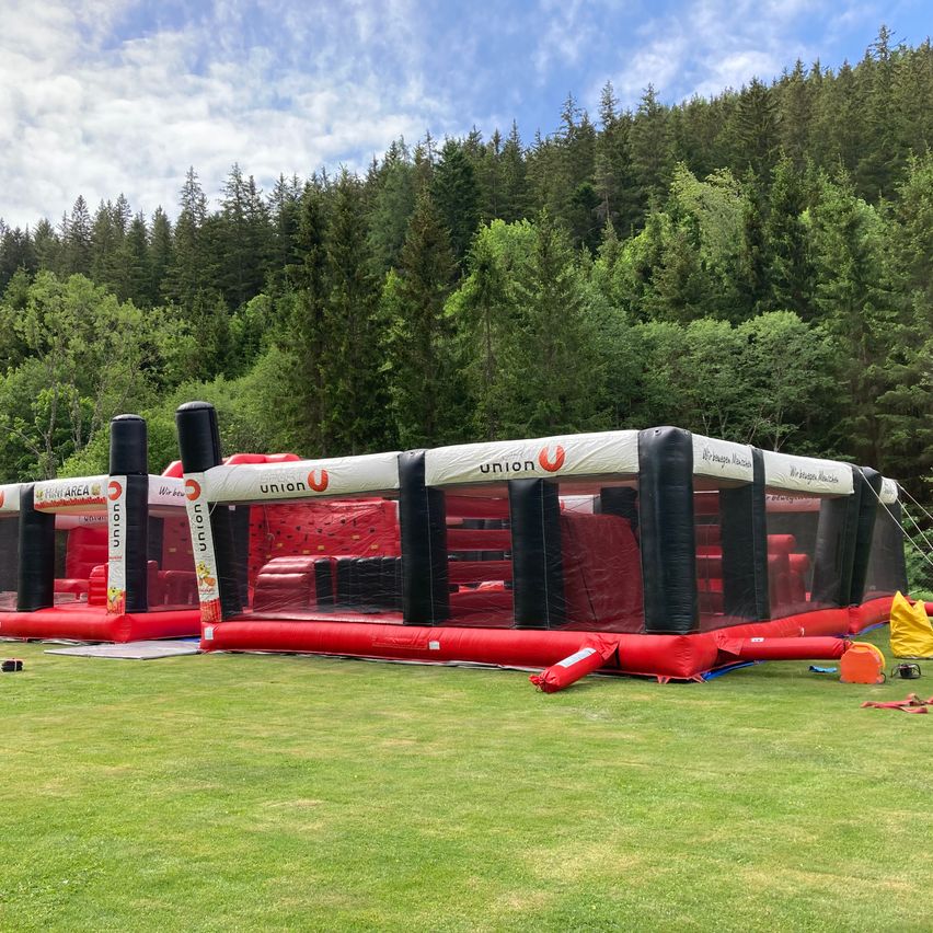 Ein großer roter und schwarzer aufblasbarer Hindernisparcours steht auf einer Wiese mit einem Wald im Hintergrund.