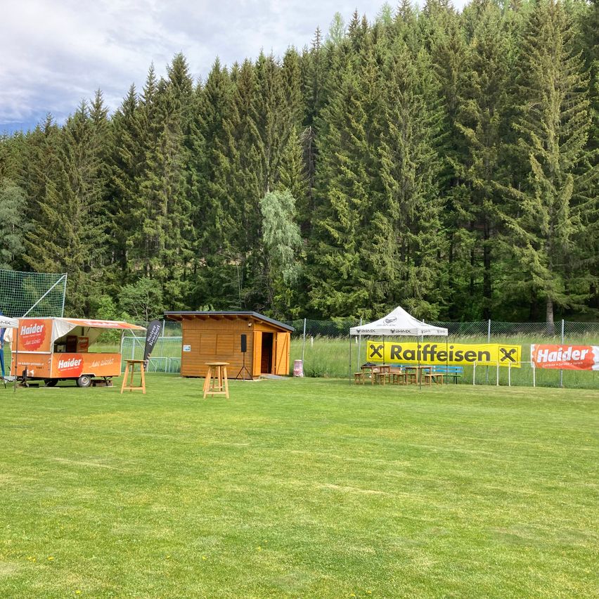 Ein breites grünes Feld mit einem kleinen Holzhütte und einem Food Truck, beide werben für Haider. Ein weißes Zelt ist unter einem Raiffeisen-Banner aufgestellt. Hohe Bäume säumen den Hintergrund.