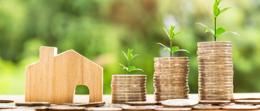 Ein Holzmodell eines Hauses mit einer kleinen Pflanze steht neben Geldstapel in zwei Töpfen. Der Hintergrund ist eine verschwommene grüne Landschaft.