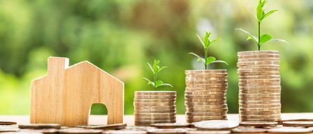 Ein Holzmodell eines Hauses, zwei Stapel Münzen mit Pflanzen und Sonnenlicht, das durch einen unscharfen grünen Hintergrund scheint.