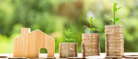 A wooden house model with a tiny plant is placed between two stacks of coins, symbolizing growth and investment.