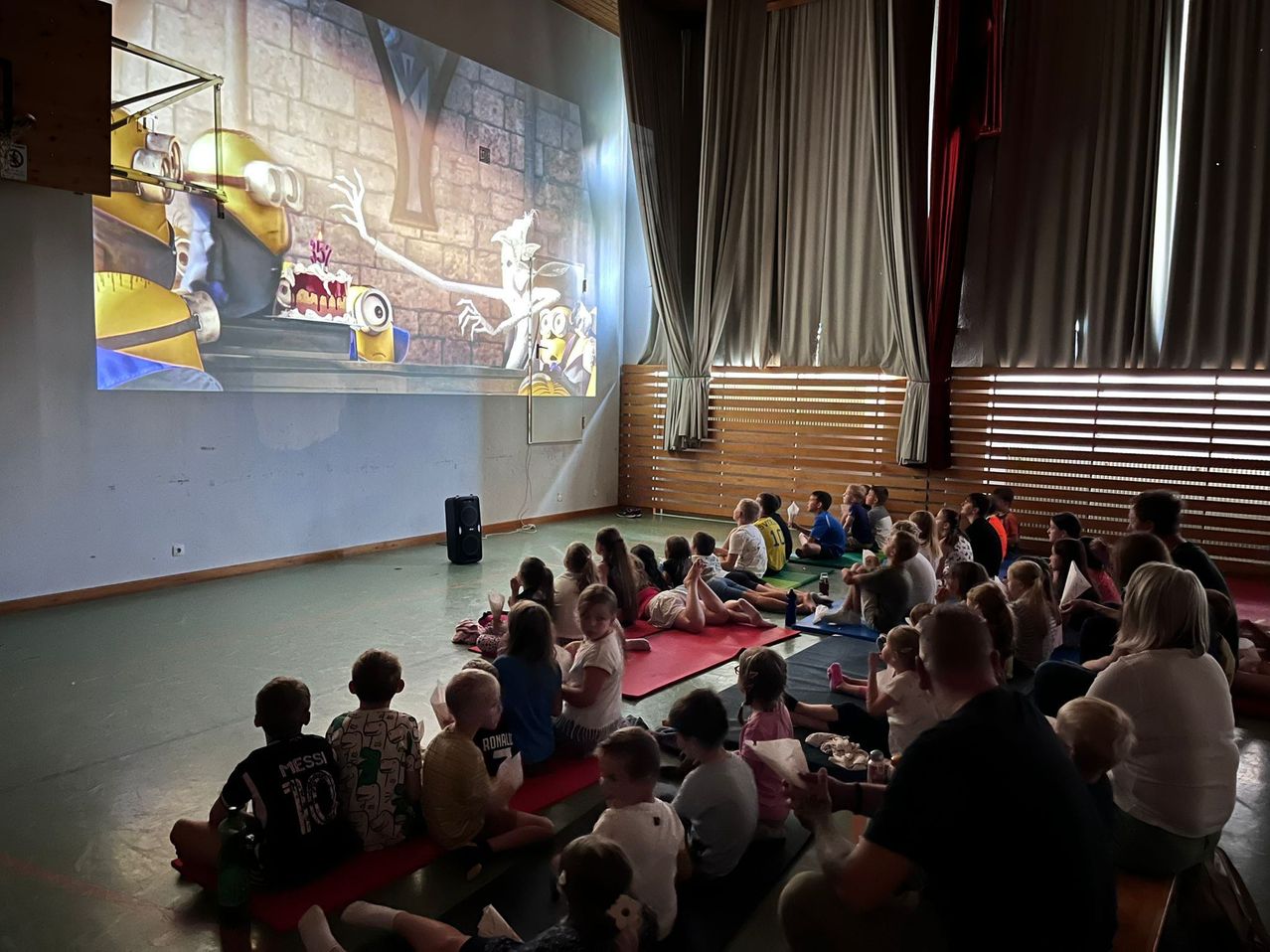 Kinder sitzen auf dem Boden und schauen einen Film auf einer großen Leinwand. Ein Lautsprecher steht auf dem Boden, und es gibt Vorhänge an der Wand.