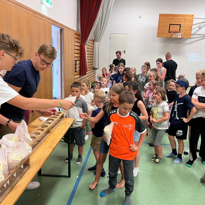 Gruppe von Kindern beobachtet einen Mann, der Popcorn von einem langen Holztisch in einer Schulsporthalle verteilt.