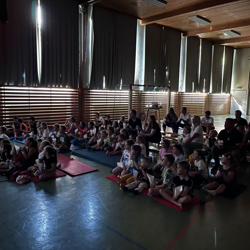 Eine Gruppe von Kindern und Erwachsenen sitzt auf roten Matten in einem Saal und schaut sich eine Präsentation an. Sie halten Tassen und Snacks. Einige Erwachsene sitzen auf Bänken. Der Raum hat Holzwände und Vorhänge.