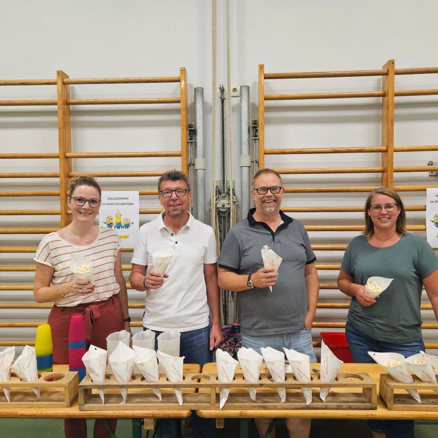 Vier Menschen stehen hinter einem Tisch mit Popcorn in Tüten und lächeln für ein Foto. Sie sind in einem Raum mit Holzwänden und einem weißen Hintergrund.