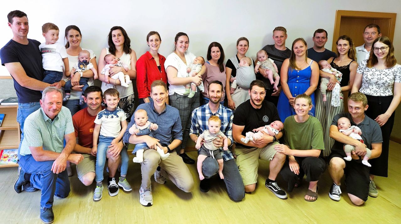 Eine Gruppe von Erwachsenen und Babys, die für ein Foto posieren, einige ihre Babys halten, andere stehen hinter ihnen, alle lächeln vor einer weißen Wand.