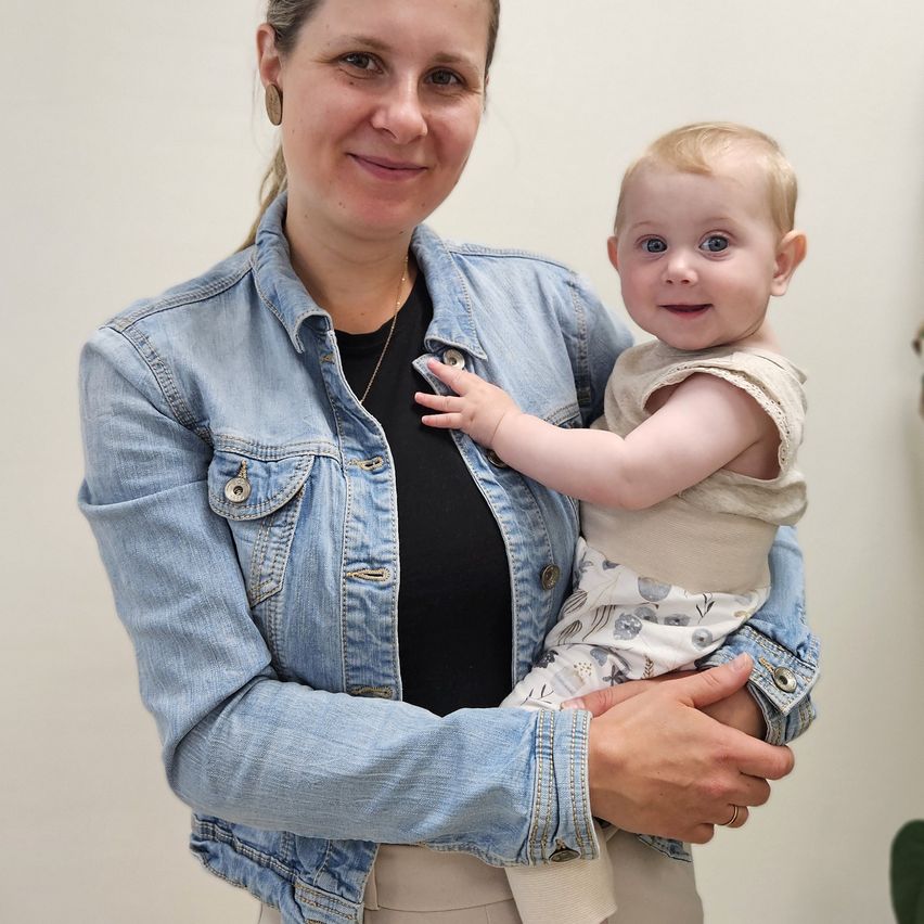 Eine Frau in einer Jeansjacke hält ein lächelndes Baby, beide schauen in die Kamera in einem weißen Raum.