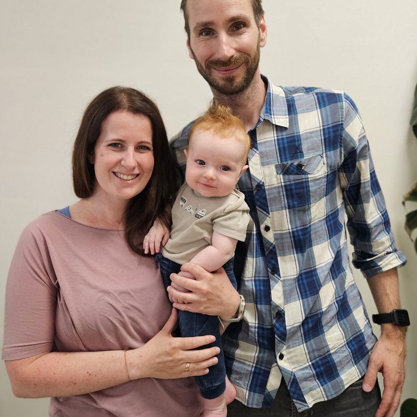 Eine Familie zu dritt, eine lächelnde Frau und ein Mann, und ein Baby, stehen eng beieinander für ein Foto. Der Mann hält das Baby, während die Frau neben ihm lächelt.