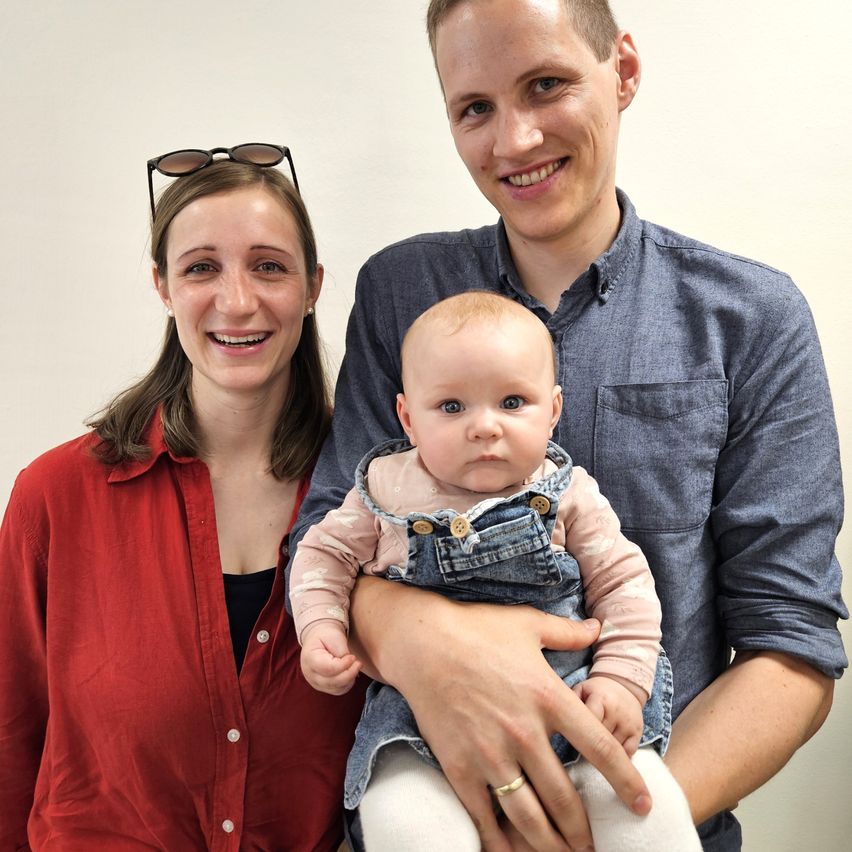 Ein Mann und eine Frau lächeln und halten ein Baby in den Armen. Das Baby trägt einen Jeans-Overall. Der Mann trägt ein blaues Hemd und die Frau ein rotes Oberteil.