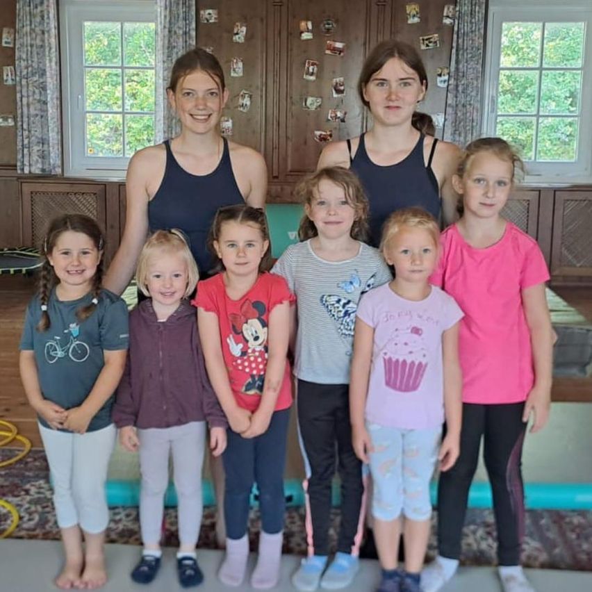 Eine Gruppe junger Mädchen und zwei Frauen stehen zusammen und lächeln für ein Foto. Die Mädchen tragen verschiedene farbige Shirts und Leggings, während die Frauen ärmellose Oberteile tragen. Hinter ihnen hat ein Raum eine Holzwand mit Bildern und einem Fenster.